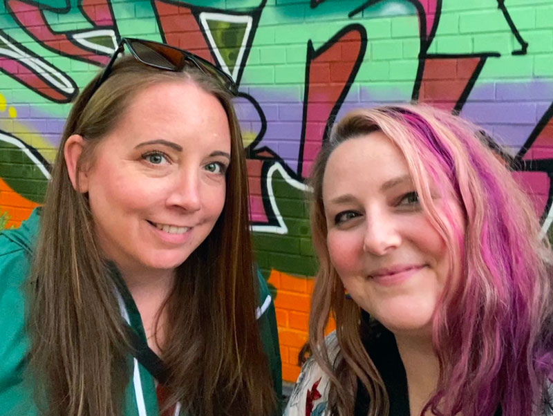 A photo of Amy and Bianca the owners of Wry and Ginger. Amy is on the left with red hair, sunglasses on her head and wearing a green jacket. Bianca is on the right with pink and blond hair.